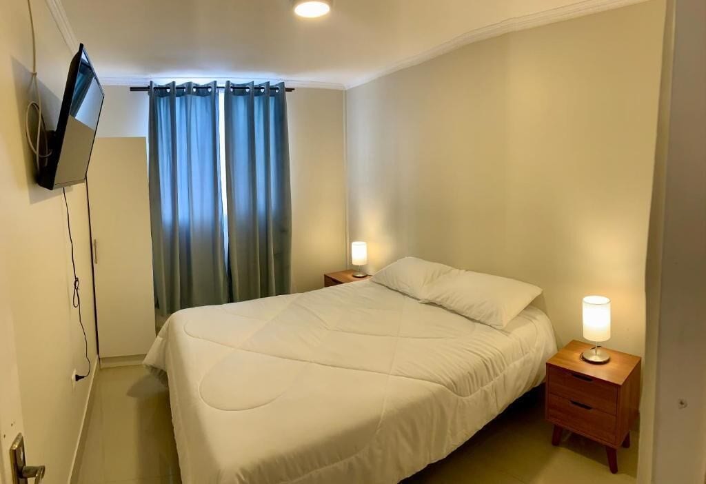 Une chambre à coucher avec un lit, deux lampes de chevet, un téléviseur fixé au mur et un rideau.