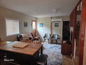 A dining area with a wooden table, chairs, and a rug. There is a door leading to another room, and a staircase is visible in the background.
