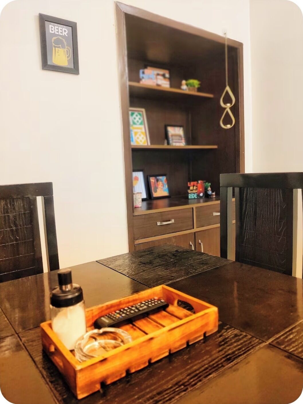 A dining table with a wooden tray holding a bottle, a remote, and a glass. Behind the table, there is a bookshelf with various items and a framed picture on the wall.