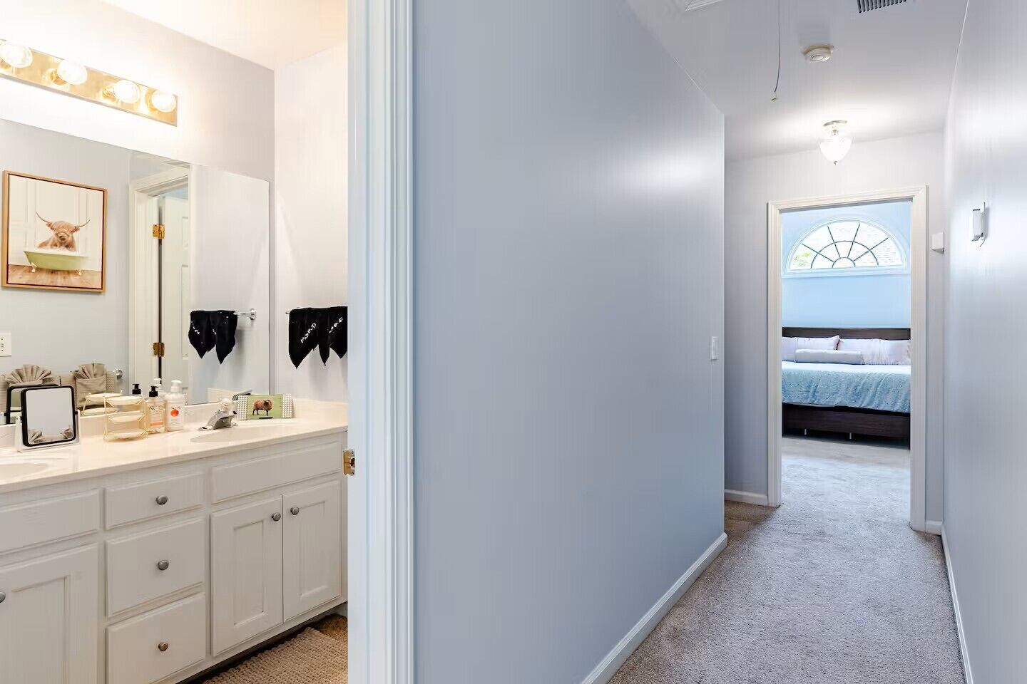 A hallway leading to a bedroom with a bed, a vanity with a mirror and lights, and a framed picture on the wall.