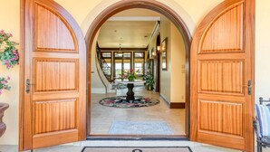 Uma entrada imponente com portas de madeira, tapete de boas-vindas e vista para um interior espaçoso com escada e mesa central.