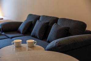Un sofá de color oscuro con varios cojines, una mesita redonda con dos tazas blancas, y una pared lisa de fondo.