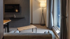 Una sala de estar moderna con un televisor de pantalla plana, un banco de madera, un sofá, una lámpara de pie y un amplio ventanal con cortinas.