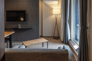 Una sala de estar moderna con un televisor de pantalla plana, un banco de madera, un sofá, una lámpara de pie y un amplio ventanal con cortinas.