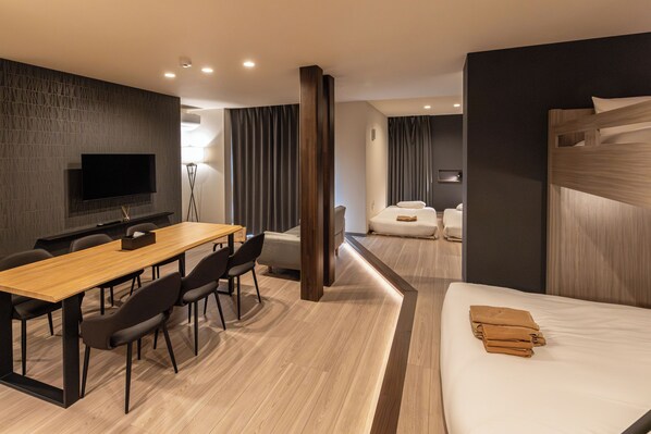 A modern hotel room with a wooden desk, chairs, a flat-screen TV, a bed with a wooden headboard, and a small seating area with a sofa. - Rela Kashiwaeki Nishi (Kashiwa)