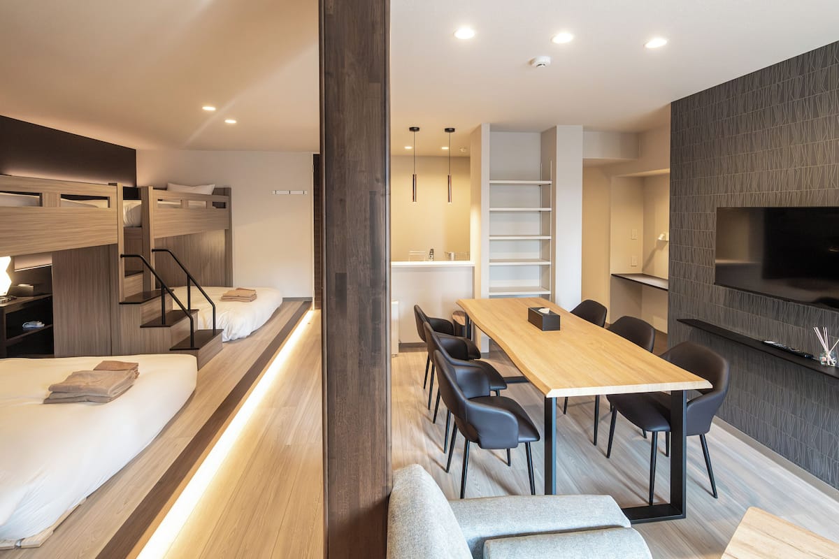 Een moderne hotelkamer met een houten bed, een zithoek met tafel en stoelen, een aan de muur bevestigde televisie en een inbouwplank.