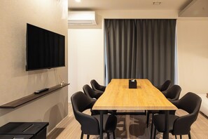 A conference room with a long wooden table, black chairs, a flat-screen TV on the wall, and a window with curtains.