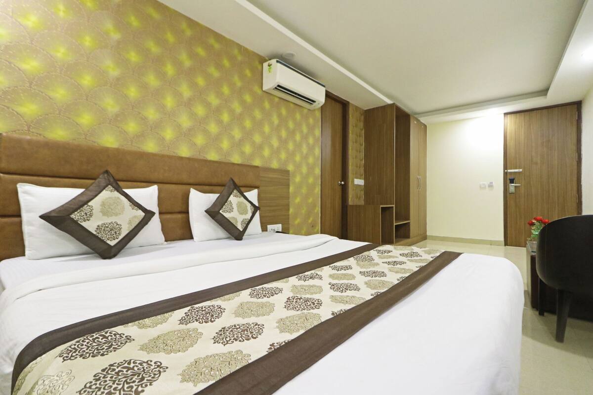 A hotel room with a large bed, a headboard with decorative pillows, an air conditioning unit, wooden wardrobes, and a desk with a chair.