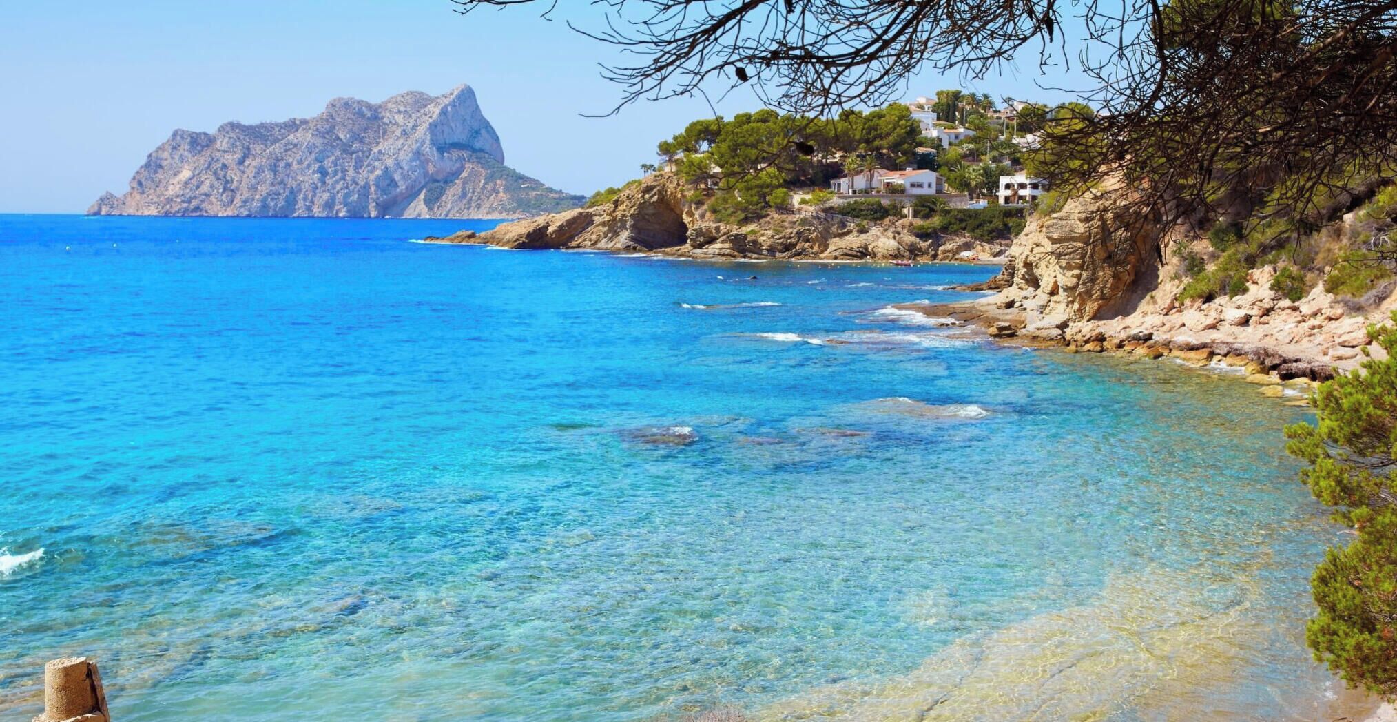 A coastal landscape with clear blue waters, rocky terrain, and a building with a sloped roof.