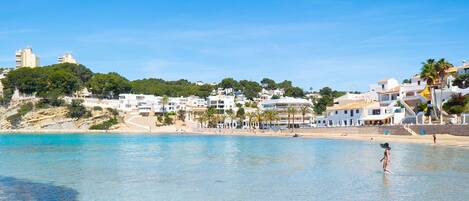 A coastal town with buildings along the beach, clear blue water, and a person standing in the water.
