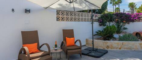 Deux fauteuils en osier avec des coussins orange, une petite table ronde avec une bouteille et des verres, et un grand parasol qui offre de l’ombre.