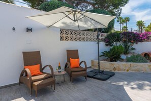 Two wicker chairs with orange cushions, a small round table with a bottle and glasses, and a large umbrella providing shade. - Villa George by Interhome (Moraira)
