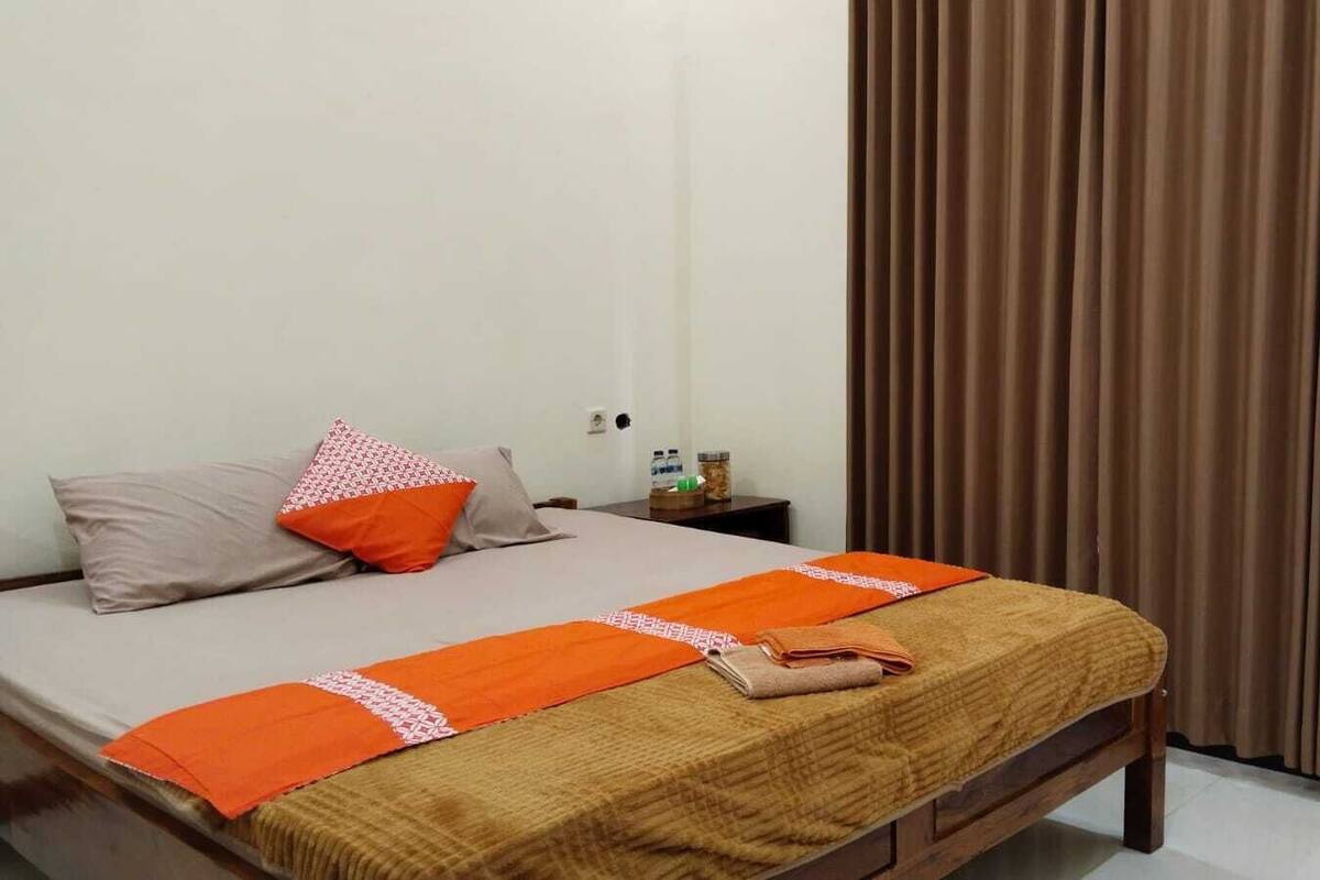 Une chambre d’hôtel avec un lit, des oreillers, une couverture, une table de chevet, un ventilateur et une fenêtre avec des rideaux.