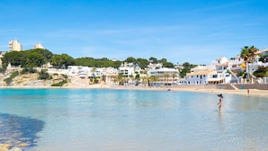 En kuststad med byggnader längs stranden, klart blått vatten och en person som står i vattnet.