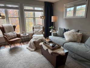 Un salon chaleureux avec un canapé, un fauteuil et un pouf, une table d’appoint avec une lampe, et une vue sur les arbres d’automne à travers de grandes fenêtres.