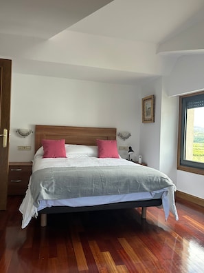 A neatly made bed with a wooden headboard, a bedside table, a nightstand, a window with a view, and a wall-mounted lamp.