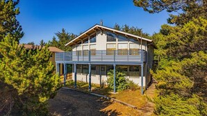 A two-story wooden cabin with a balcony, surrounded by dense pine trees. - 3BR Copalis Beach Cottage | Fireplace | Dogs OK (Copalis Beach)