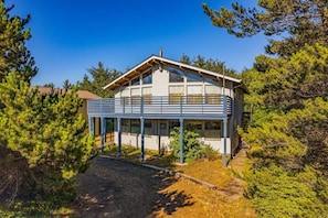 A two-story wooden cabin with a balcony, surrounded by dense pine trees.