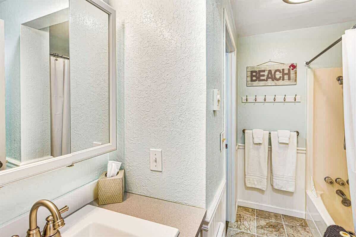 Un baño con un espejo grande, un lavamanos, una ducha y un perchero con toallas.