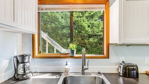 A kitchen with a stainless steel sink, a coffee maker, a toaster, and a window with a view of trees. - 3BR Copalis Beach Cottage | Fireplace | Dogs OK (Copalis Beach)