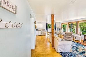 A spacious living area with a wooden floor, a sofa, a chair, and a coffee table. There is a kitchen in the background with a microwave and cabinets.