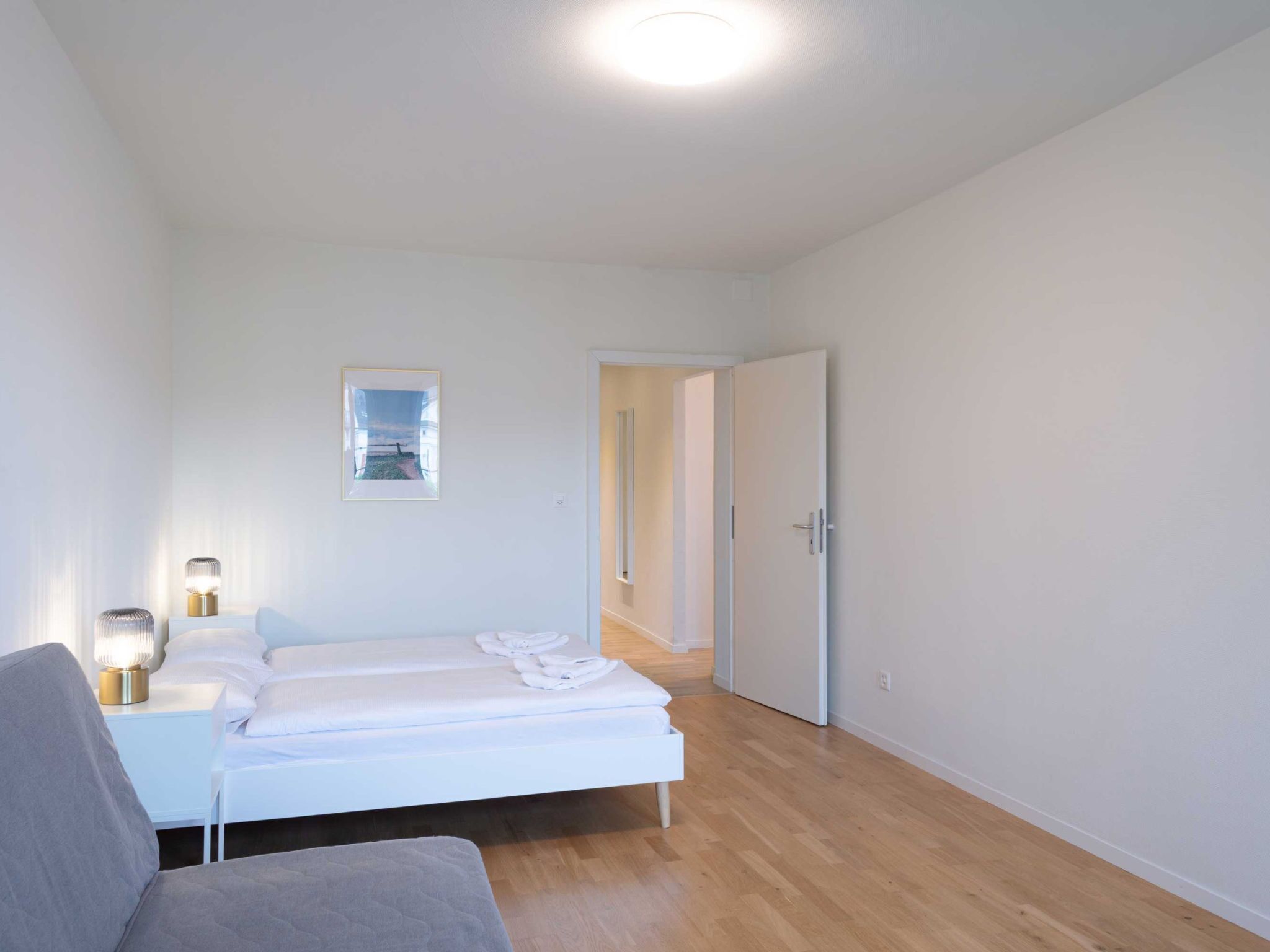 Un dormitorio con cama, mesitas de noche, una silla, una puerta y un pasillo.