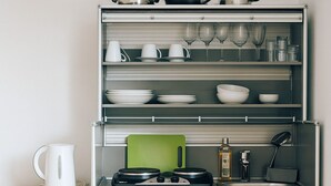 A kitchen with a built-in storage unit, a kettle, a cutting board, a sink, and a wine glass rack.