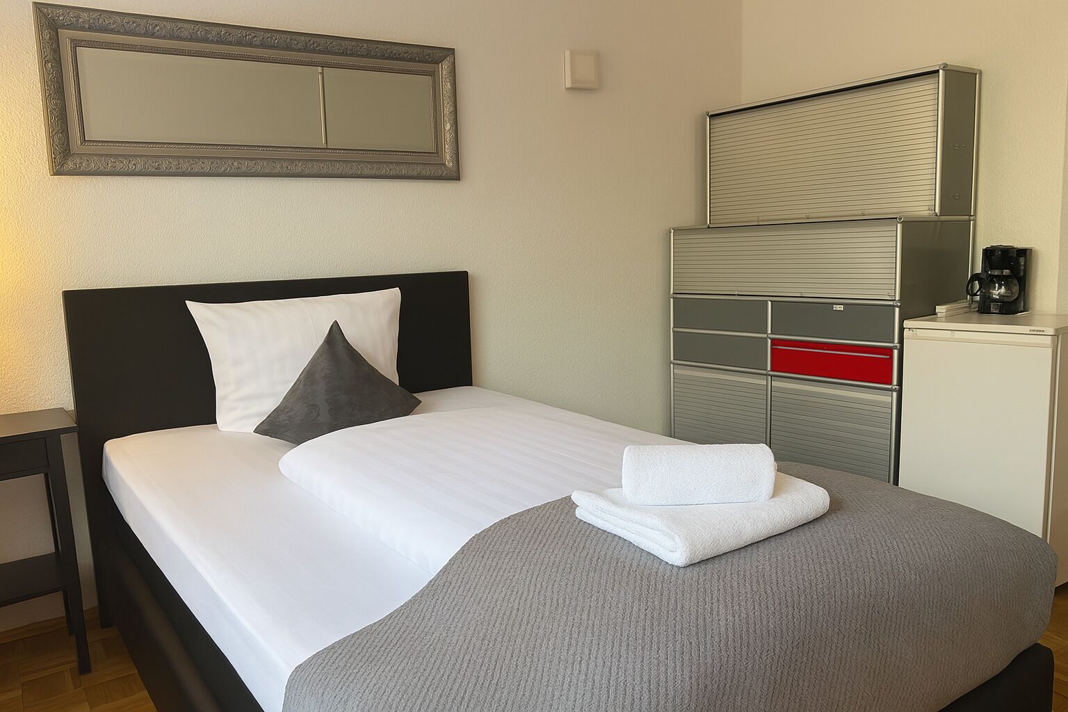 A neatly made bed with pillows, a folded towel, a bedside table, a mirror, and a minibar with a coffee machine.