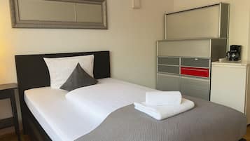 A neatly made bed with pillows, a folded towel, a bedside table, a mirror, and a minibar with a coffee machine.