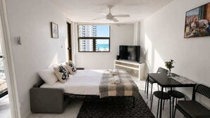 A bedroom with a bed, a TV, a desk with chairs, a window with a view, and a ceiling fan. - Beachside 1BR Retreat | Ocean Views, Wi-Fi & Pool (Surfers Paradise)