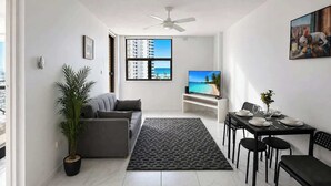 A modern living room with a sofa, a dining table set for four, a large window with a view of buildings and a beach, and a ceiling fan. - Beachside 1BR Retreat | Ocean Views, Wi-Fi & Pool (Surfers Paradise)