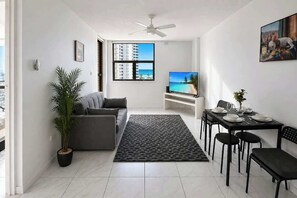 Ein modernes Wohnzimmer mit einer Couch, einem Esstisch für vier Personen, einem großen Fenster mit Blick auf Gebäude und einen Strand sowie einem Deckenventilator.