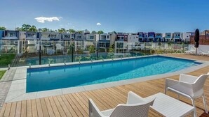 A swimming pool with a wooden deck, white lounge chairs, and modern buildings in the background. - Luxury Lakeview 3BR Escape (Robina)