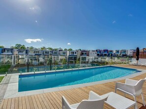 A swimming pool with a wooden deck, white lounge chairs, and modern buildings in the background. - Luxury Lakeview 3BR Escape (Robina)