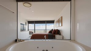 A modern bathroom with a large bathtub, a visible bedroom with a bed and window, and a view of the sea. - L54 Sky Ocean View Apt Heated Pool (Surfers Paradise)