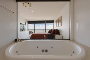 A modern bathroom with a large bathtub, a visible bedroom with a bed and window, and a view of the sea. - L54 Sky Ocean View Apt Heated Pool (Surfers Paradise)