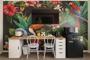 A room with a flat-screen TV mounted on the wall, a wooden desk with two white chairs, a black refrigerator, and a wall mural with tropical flowers and a parrot. - L22 Ocean View 2 Beds Wi-Fi Netflix (Surfers Paradise)
