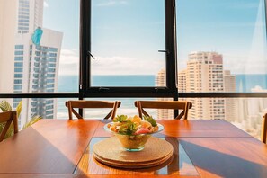 A dining table with chairs and a bowl of fruit in a room with a large window overlooking a cityscape. - Level 29 Sky 2BR Ocean View Apartment (Surfers Paradise)