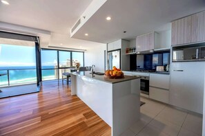 A modern kitchen with a large island, a dining area with a view of the ocean, and a spacious living area with wooden flooring. - Level 29 Sky 2BR Ocean View Apartment (Surfers Paradise)
