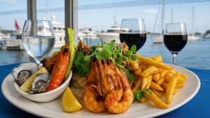 Eine Platte mit Meeresfrüchten, darunter Garnelen, Austern und Pommes, begleitet von Weingläsern und Wassergläsern, vor einer Bootsanlegestelle.