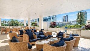 A rooftop lounge with wicker furniture, glass tables, and a view of city buildings. - Surfers 1BR Retreat: Pool, Spa, Gym & Fast Wi-Fi (Surfers Paradise)
