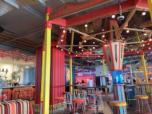 Een levendige bar met kleurrijke krukken, een toog en een plafond met zichtbare balken en hangende lampen.