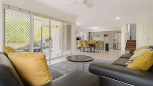 A modern living room with a sofa, a coffee table, and a dining area with chairs. - Dreamworld Home with Pool, BBQ, WI-FI and Backyard (Oxenford)