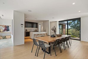 A modern kitchen with a central island, a dining area with a large wooden table and chairs, and a view through large windows.