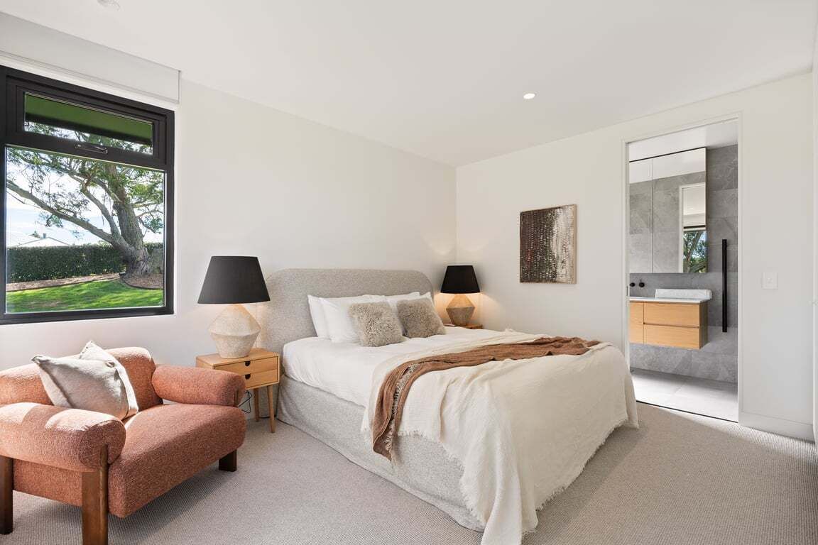 A modern bedroom with a large bed, bedside lamps, a nightstand, a chair, a window with a view, and a bathroom with a mirror and sink.
