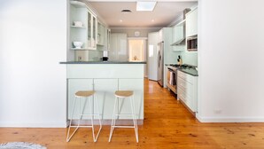 A modern kitchen with a central island, bar stools, and a view into the dining area. - Casaba Villa - Your Beachside Garden Oasis (Glenelg South)