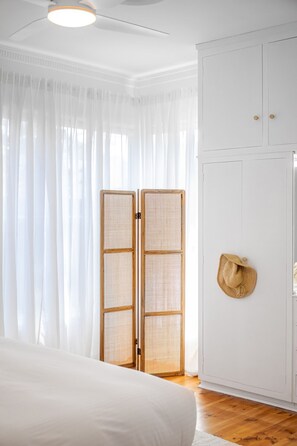 A bedroom with a bed, white curtains, a woven screen door, and a straw hat on the wall. - Casaba Villa - Your Beachside Garden Oasis (Glenelg South)