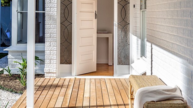 A wooden deck with a bench and a door leading to a room with a white wall and a textured pattern. - Casaba Villa - Your Beachside Garden Oasis (Glenelg South)