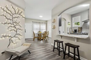 Una cocina moderna con encimera blanca, taburetes oscuros y una zona de comedor con sillas de madera.