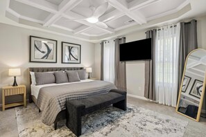 Un dormitorio con cama, mesitas de noche, un banco, ventilador de techo, televisor y cortinas.